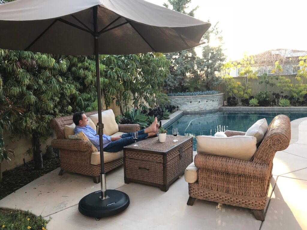 A man is sitting on outdoor patio furniture by the pool relaxing under an umbrella